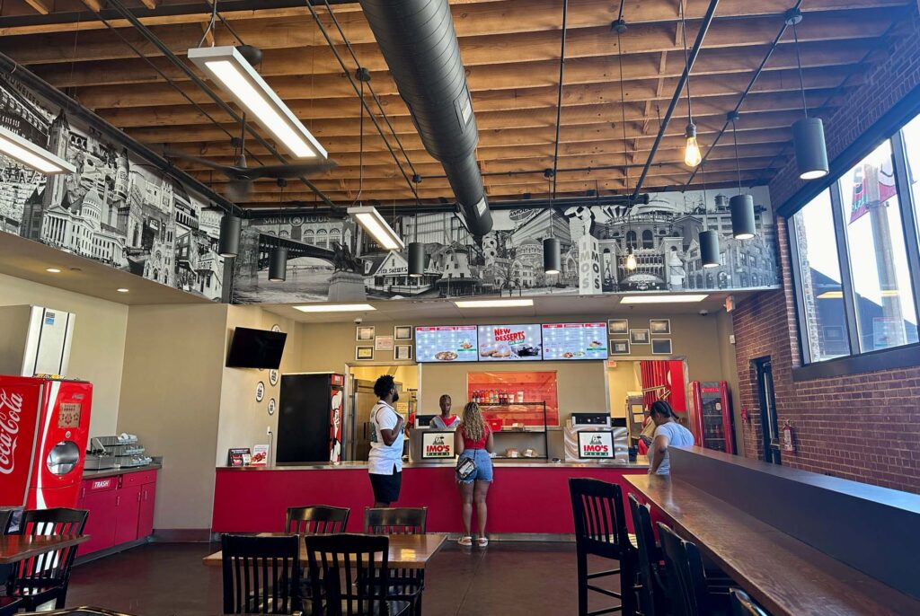 Interior of an Imo's Pizza with customers ordering at a red counter, menu screens, and St. Louis mural along the wall