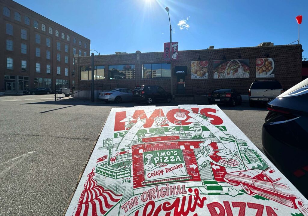 An Imo's Pizza box with St. Louis-themed artwork sits on a car outside the brick Imo's restaurant under a sunny blue sky