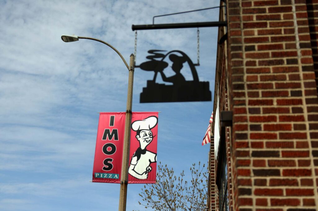 Red Imo's Pizza banner with chef logo on a lamppost beside a brick building and hanging silhouette sign against blue sky
