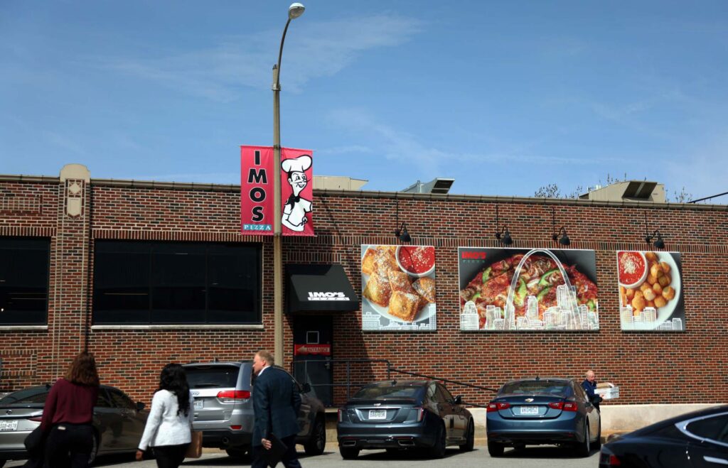 Imo's Pizza brick storefront with food posters and banner, cars parked out front and people walking by