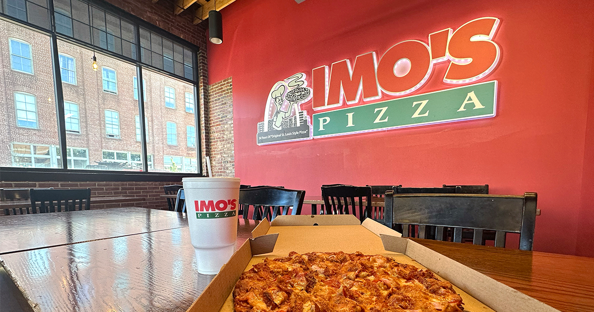 A pizza in a box and a drink cup on a table inside an Imo's Pizza restaurant with a red neon logo wall