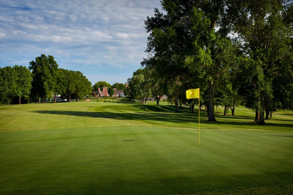 Yellow flag on a manicured golf green lined with tall trees, brick clubhouse in the distance under a blue sky