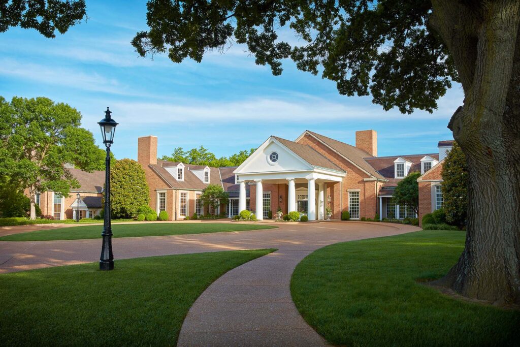 Elegant brick estate with white columned portico entrance, manicured lawn, lamppost, and curved walkway under a blue sky