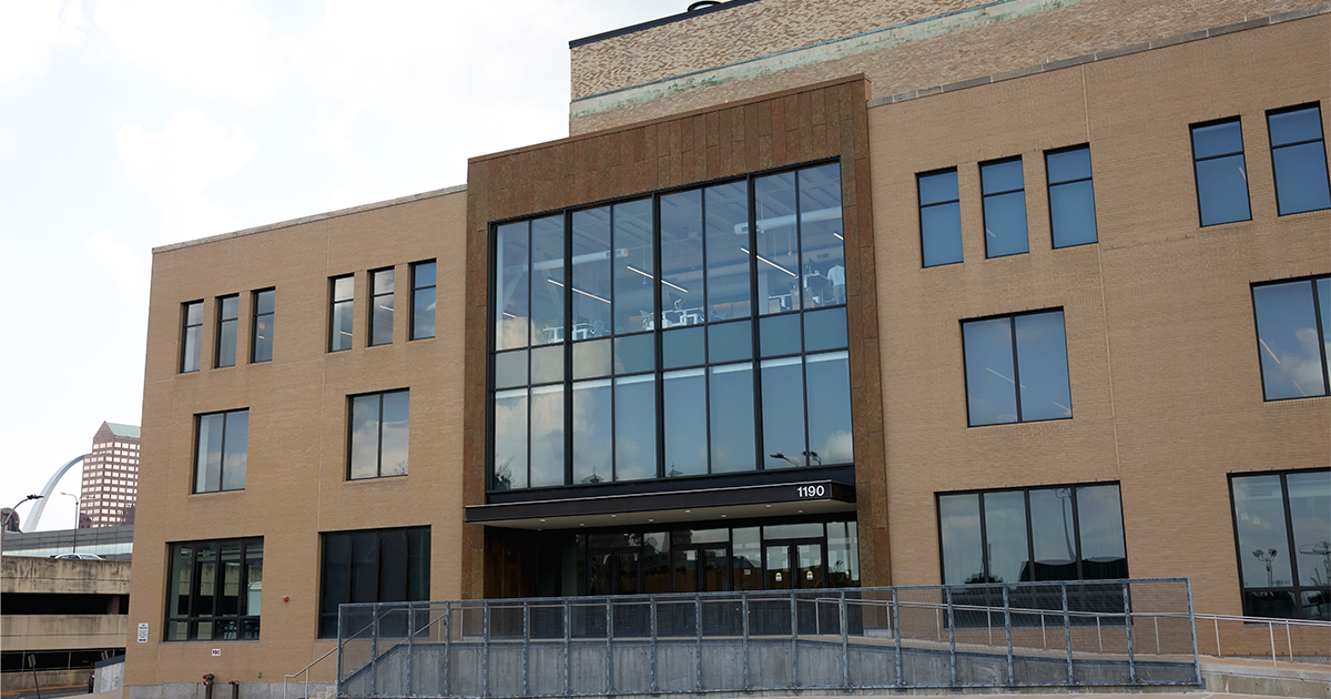 Modern tan brick office building numbered 1190 with large glass windows and a ramped entrance, Gateway Arch visible in the distance