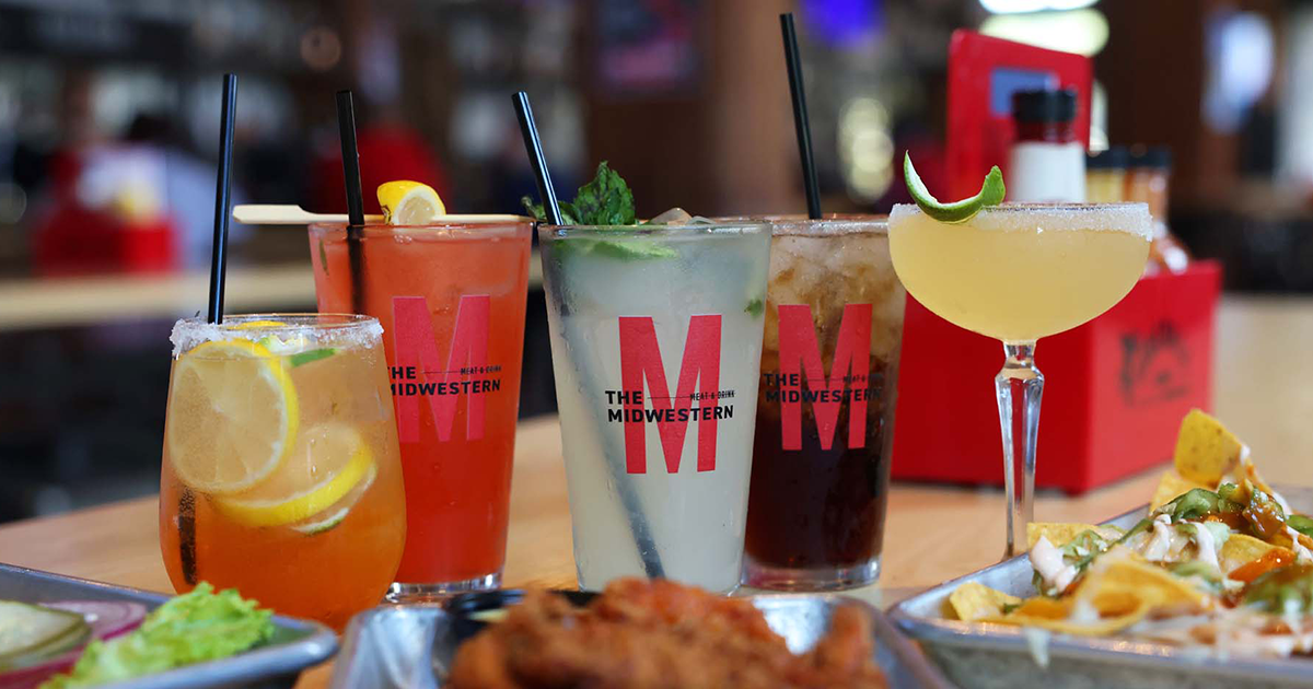 Assorted cocktails in glasses branded "The Midwestern" beside plates of nachos and wings