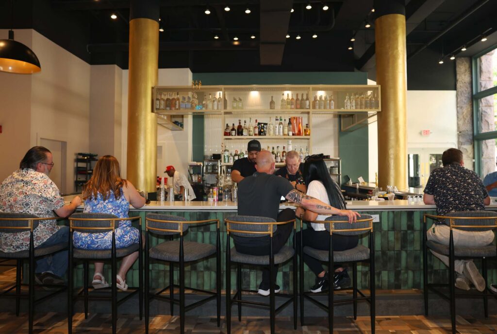 Patrons on stools at a green-tiled bar with bartenders serving and shelves of liquor bottles behind