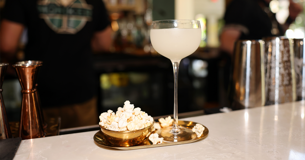 Pale cocktail in a coupe glass with a gold bowl of popcorn on a bar counter, bartender in the background