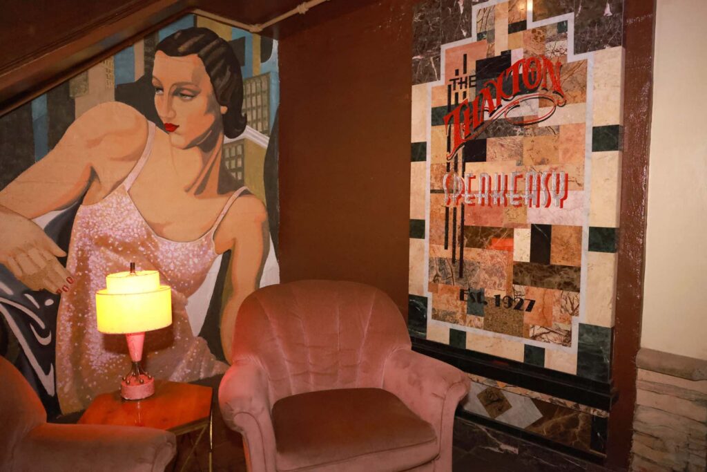 Art Deco lounge with a 1920s woman mural, pink velvet chair, lit lamp, and marble sign reading The Thaxton Speakeasy Est. 1927