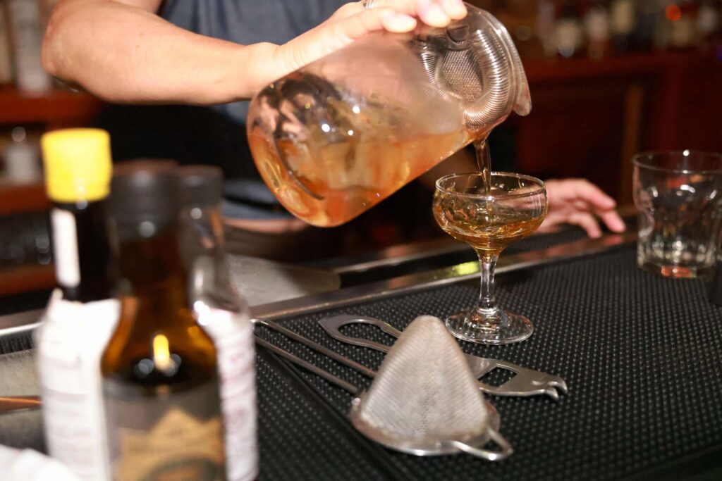 Bartender straining an amber cocktail from a mixing glass into a coupe on a bar mat