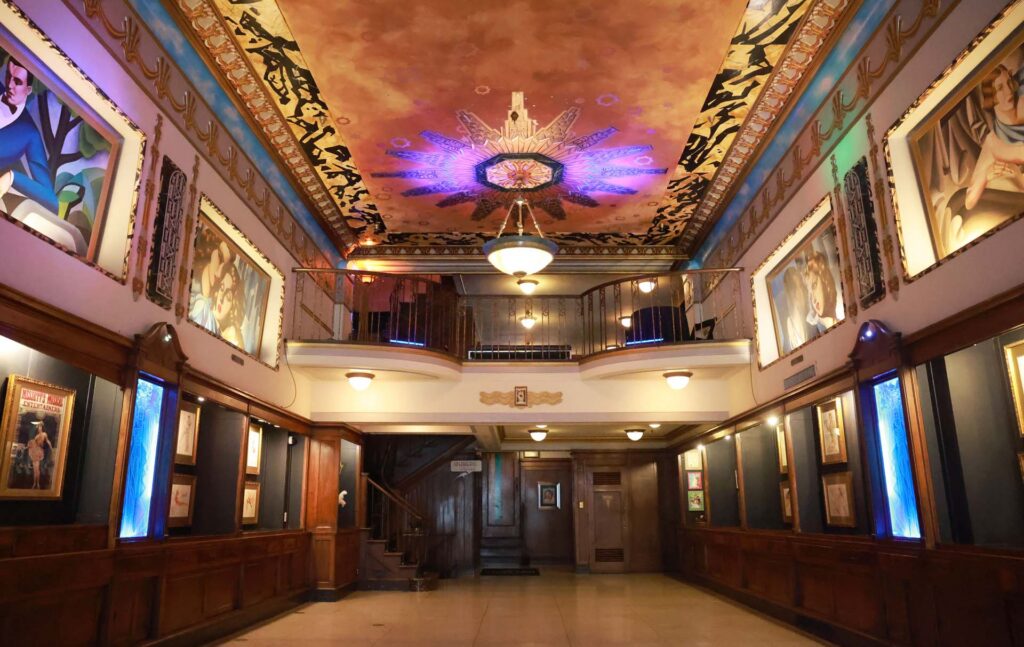 Ornate Art Deco lobby with painted ceiling, glowing star chandelier, wood paneling, and framed vintage artwork on the walls