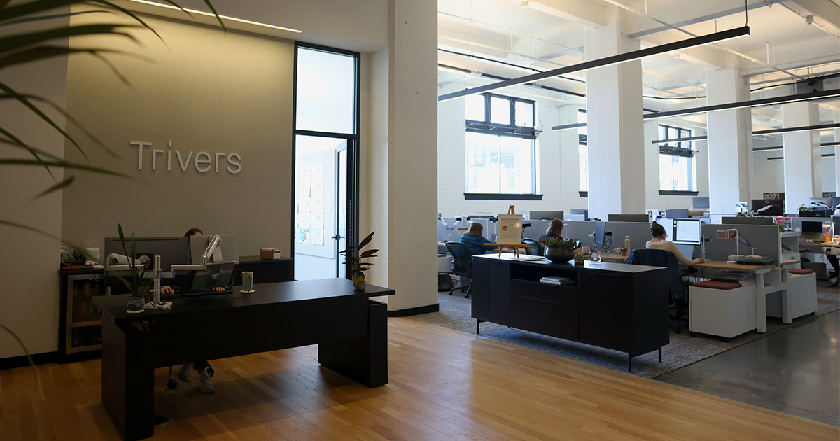 Modern open-plan office with Trivers signage on the wall, dark desks, and employees working at workstations