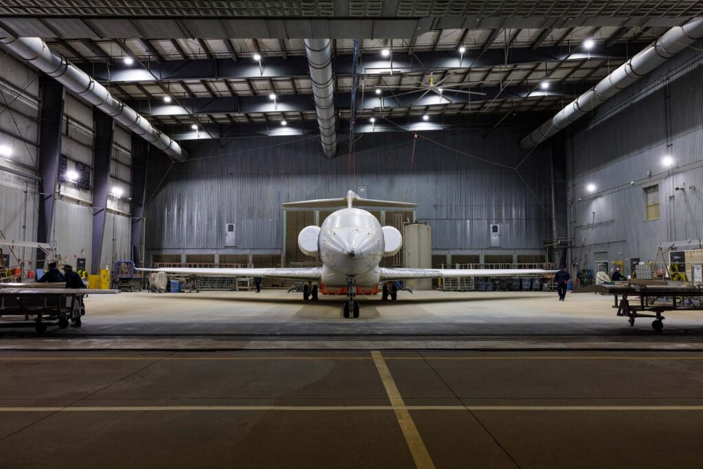 A private jet wrapped in protective covering sits in a large maintenance hangar as workers move nearby