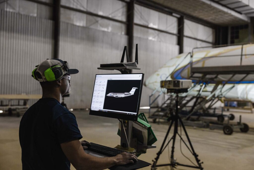 Worker with ear protection views a G600 jet design on a monitor in a hangar, with an aircraft fuselage and projector nearby