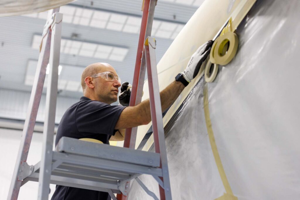 Worker on a ladder applying masking tape and protective film to an aircraft fuselage in a hangar