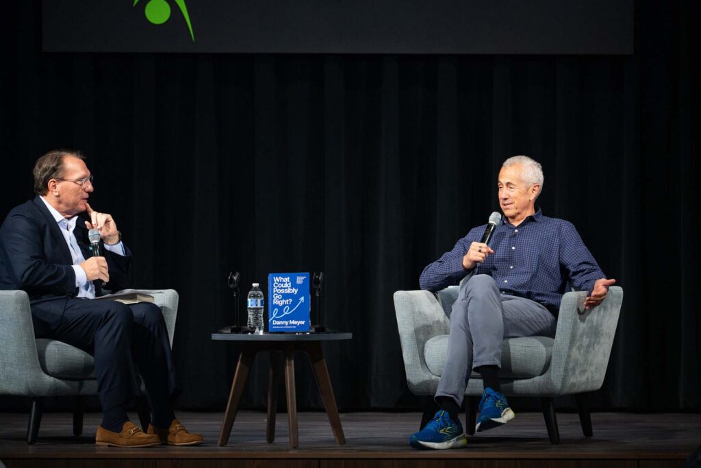 Two men in armchairs with microphones in an onstage conversation beside a table displaying Danny Meyer's book "What Could Possibly Go Right?