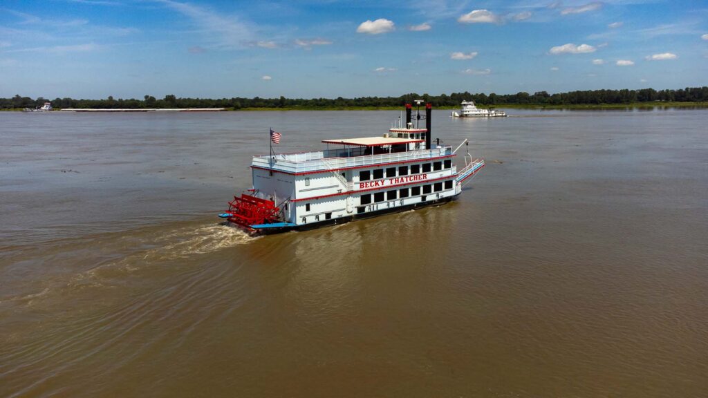 Paddle steamer "Becky Thatcher" cruising a wide muddy river, with a towboat and barge in the background