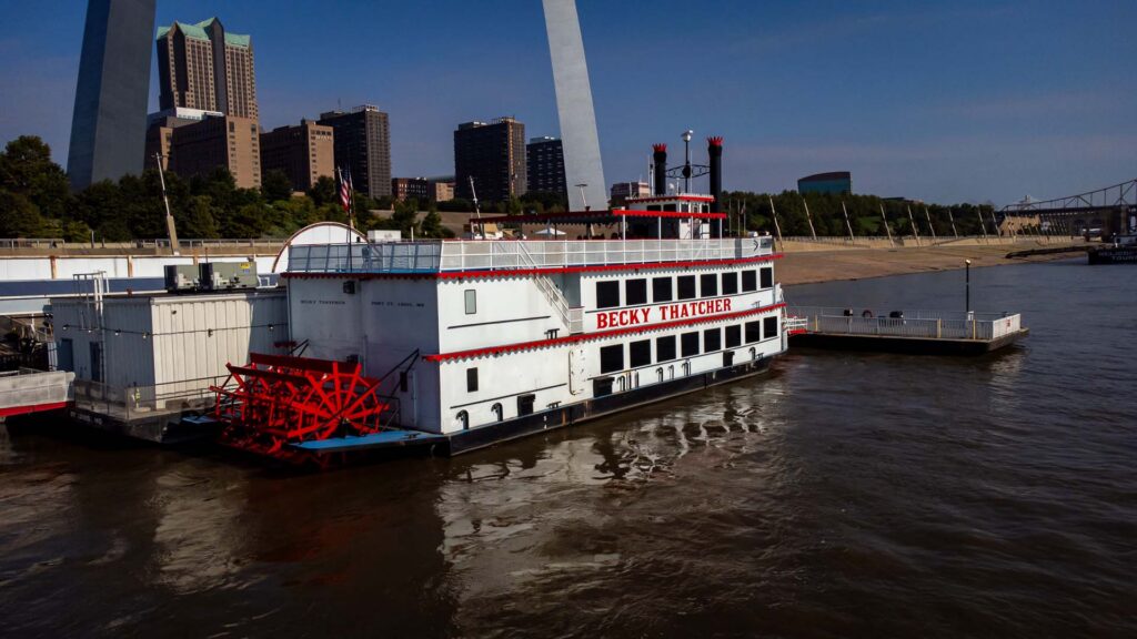 The "Becky Thatcher" paddlewheel riverboat docked on the Mississippi River with the Gateway Arch and St. Louis skyline behind