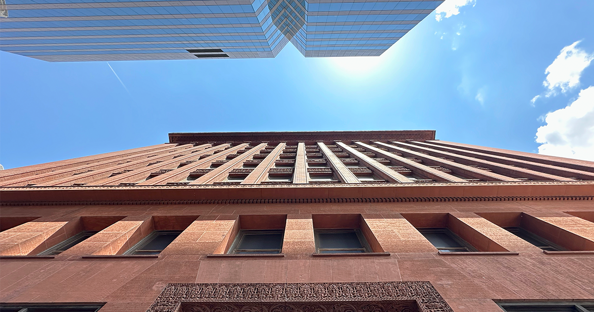 Upward view of an ornate red-brown masonry building beside a modern glass skyscraper against a bright blue sky