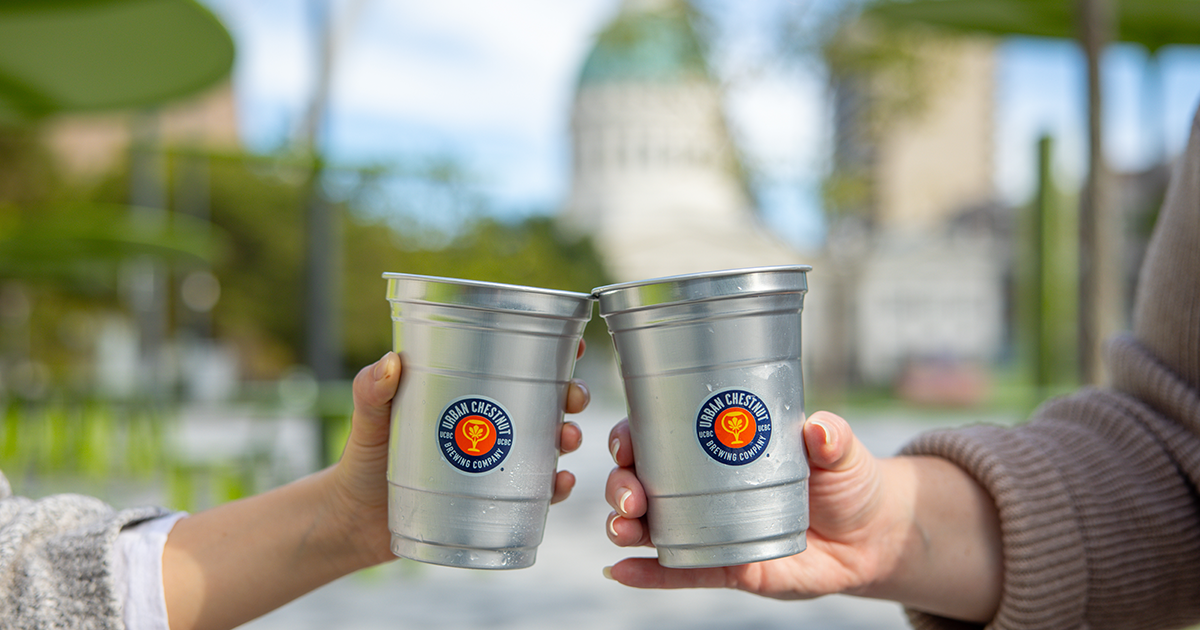 Two hands toasting with metal Urban Chestnut Brewing Company cups outdoors near a domed building