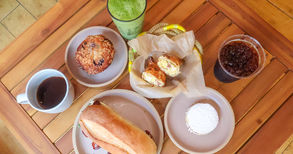 Breakfast spread on a wooden table with a muffin, pastries, bread, powdered pastry, coffee, matcha, and iced tea