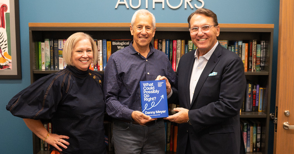 danny meyer featured photo Three people smiling in front of a bookshelf, holding Danny Meyer's book "What Could Possibly Go Right?