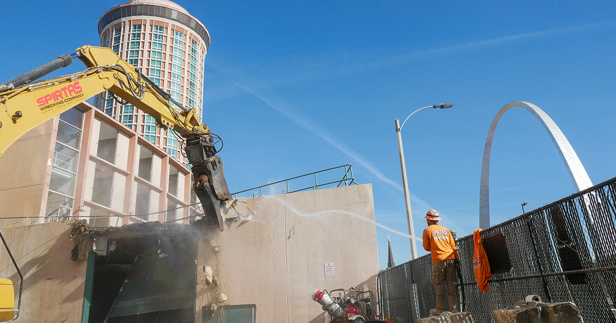 Spirtas Wrecking Company excavator demolishes a building as a worker sprays water for dust control, with the Gateway Arch in the background