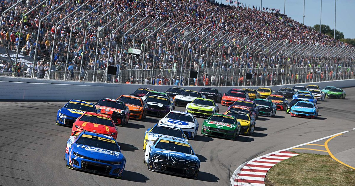 A packed field of NASCAR stock cars races through a turn before crowded grandstands