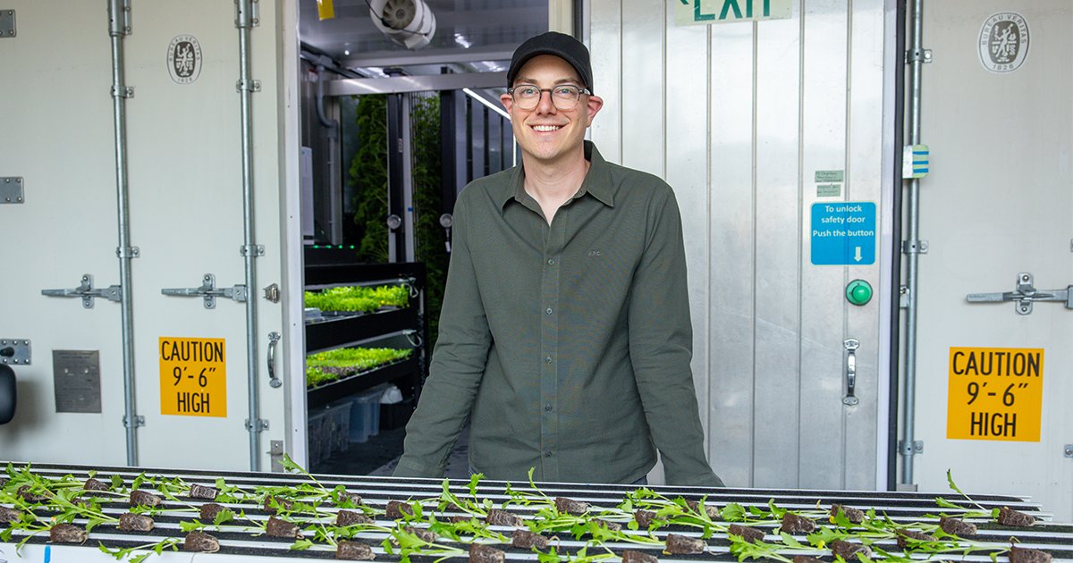 Smiling man in a cap stands behind seedling trays at the entrance of a container hydroponic farm