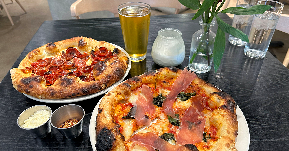 Two wood-fired pizzas, one pepperoni and one with prosciutto and basil, with beer, water, and condiments on a dark table