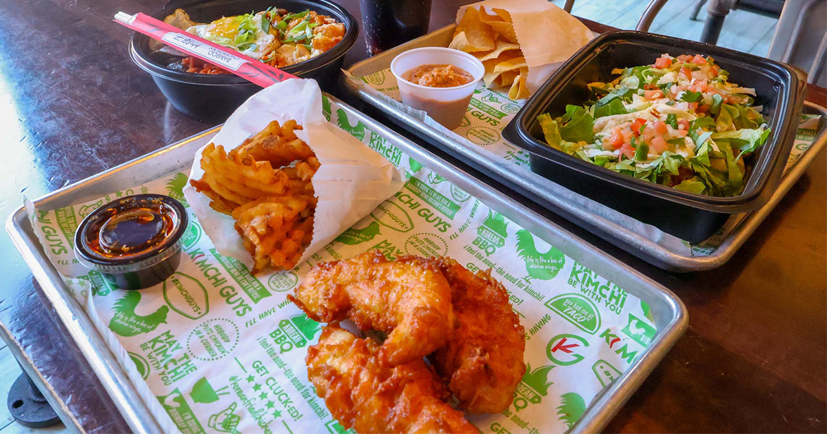 Trays of food including fried chicken, waffle fries, a salad bowl, and dipping sauces on Kimchi Guys branded paper